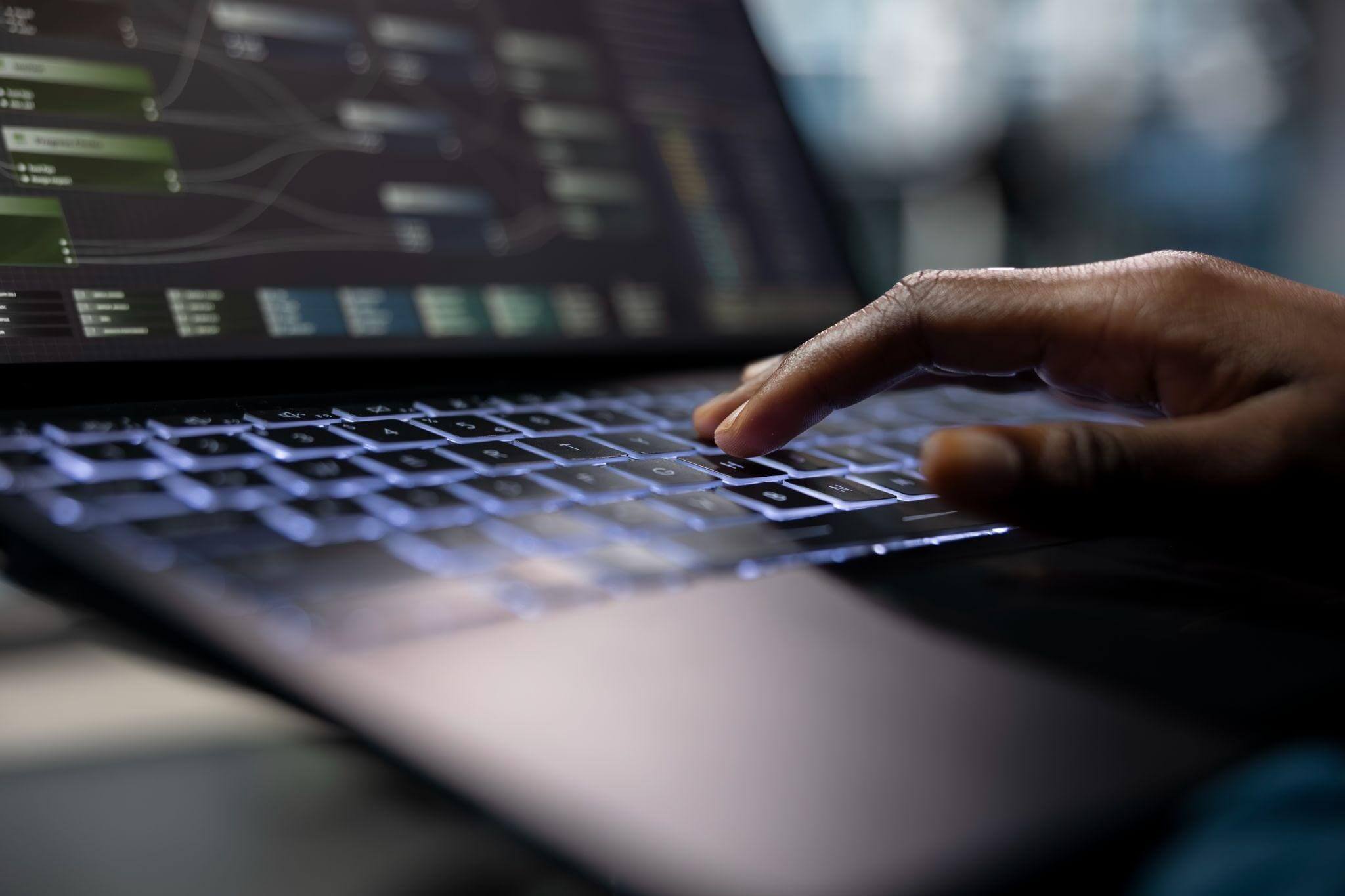 Close-up lateral da mão de uma pessoa com pele escura digitando em um teclado iluminado de um laptop, em um ambiente escuro ou de pouca luz.