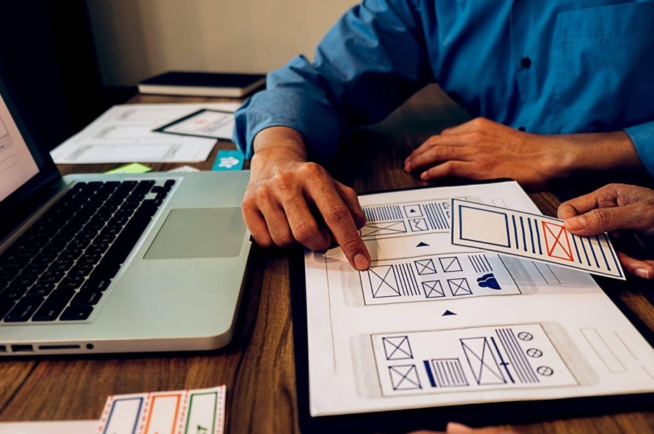 Equipe de desenvolvedores analisando wireframes em papel sobre uma mesa de madeira, focando no planejamento da interface para um redesign de site em 2026.