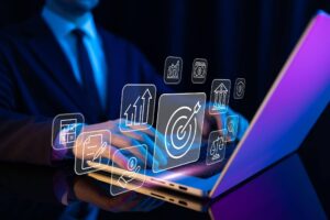 Close-up das mãos de um profissional em um notebook, com diversos ícones digitais flutuando sobre o teclado, como gráficos de crescimento, alvos e documentos. A iluminação é azul e moderna, simbolizando o planejamento estratégico por trás de o que é MVP e a validação de ideias de negócio.